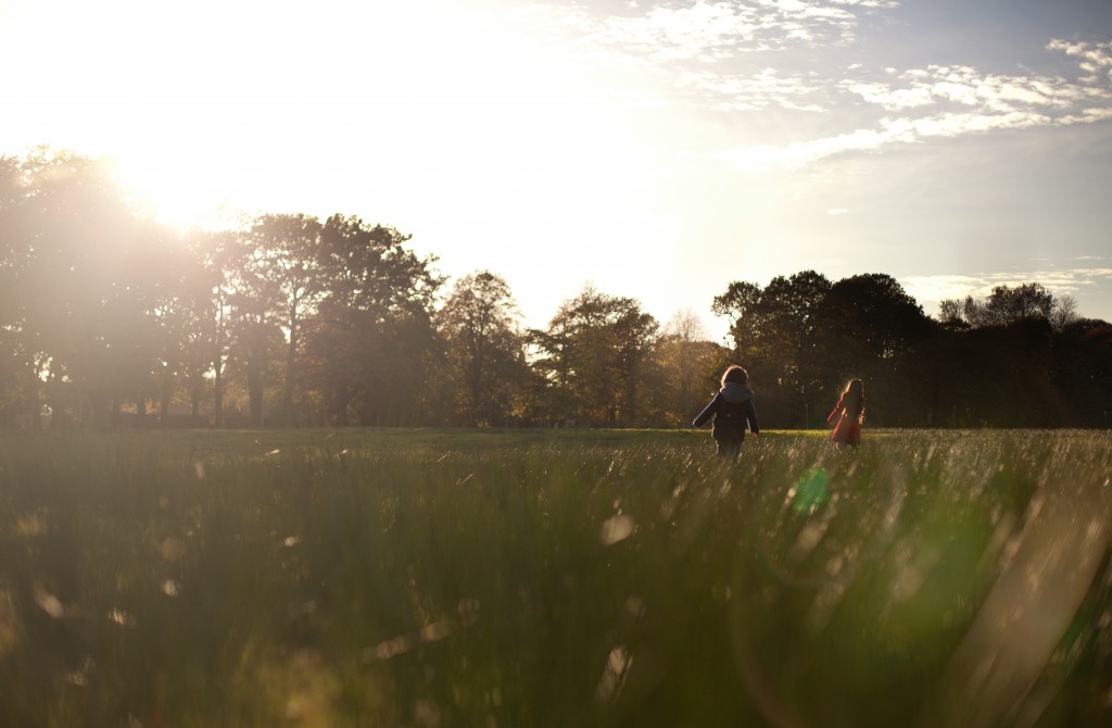 Running in the sun, two siblings charge across a field in Ashton Park, Preston 